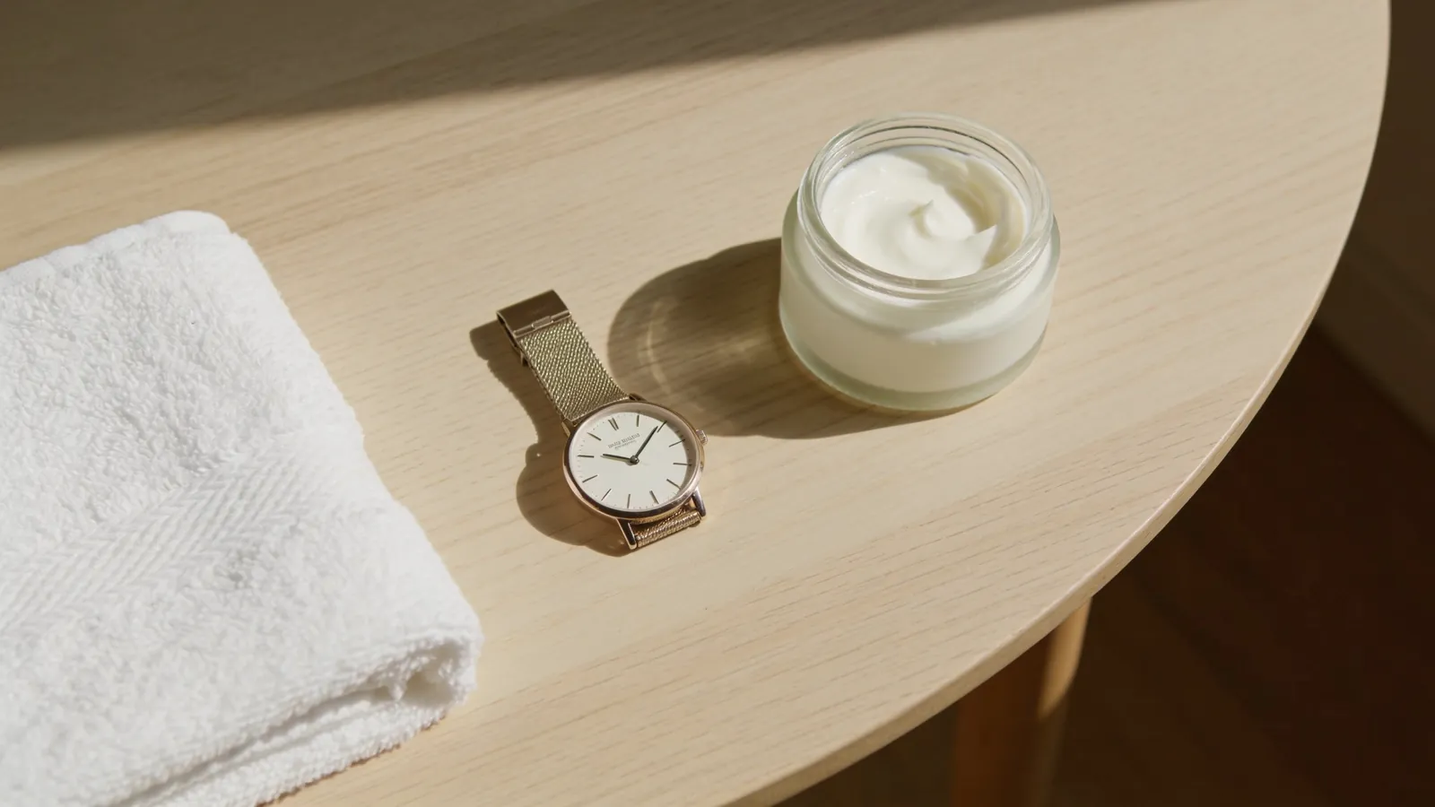 Wristwatch, folded towel and cream jar on a light wooden table