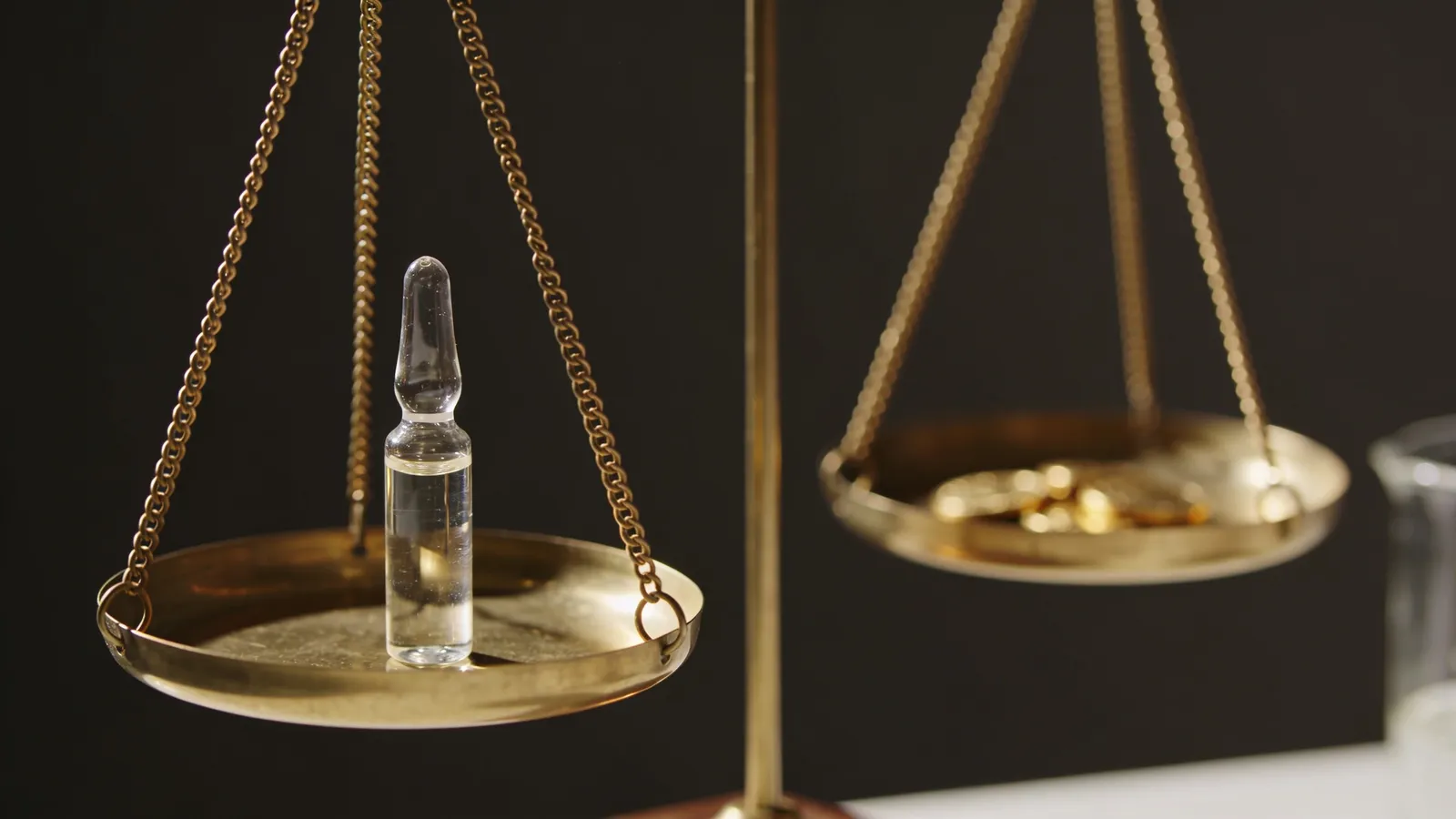 Brass balance scale weighing a serum ampoule against gold coins, illustrating treatment prices